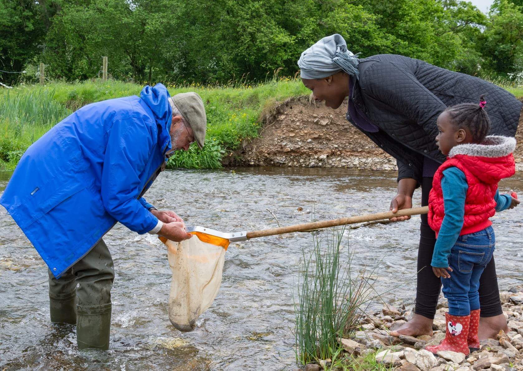 A woman standing with toddler stands by a stream with fishing net. An older man who is wading in the stream is examining the contents of the net.