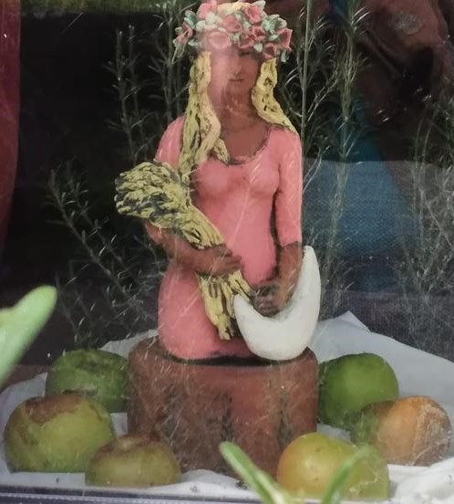 A figurine of a woman in a pink dress and flowery headdress holding a wheat sheaf and surrounded by fresh apples